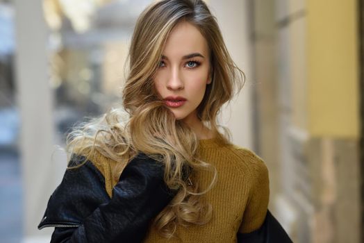 Blonde woman in urban background. Beautiful young girl wearing black leather jacket and mini skirt standing in the street. Pretty russian female with long wavy hair hairstyle and blue eyes.