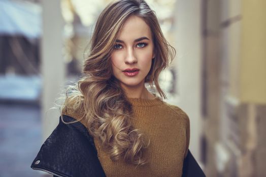 Blonde woman in urban background. Beautiful young girl wearing black leather jacket and mini skirt standing in the street. Pretty russian female with long wavy hair hairstyle and blue eyes.