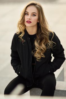 Young blonde girl with beautiful blue eyes wearing black jacket and trousers outdoors. Pretty russian female with long wavy hair hairstyle. Woman in urban background.