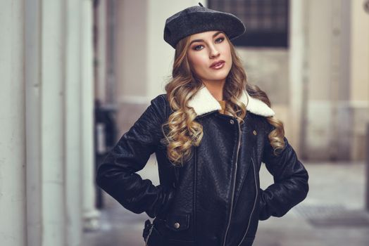 Blonde russian woman in urban background. Beautiful young girl wearing beret, black leather jacket and mini skirt standing in the street. Pretty female with long wavy hair hairstyle and blue eyes.