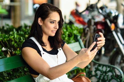 Young woman taking photographs with her smart phone outdoors. Girl sitting in urban bench. Technology concept.
