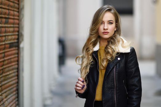Blonde woman in urban background. Beautiful young girl wearing black leather jacket and mini skirt standing in the street. Pretty russian female with long wavy hair hairstyle and blue eyes.