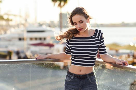 Blonde woman, model of fashion, standing in a harbor with boats and sea in the background. Beautiful young girl wearing striped t-shirt and blue jeans. Pretty russian female with pigtail.