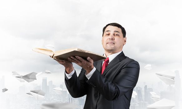 Serious businessman holding open book. Man in business suit standing on cloudy cityscape background with flying paper planes. Lawyer reading legal regulation book. Business accounting and consulting