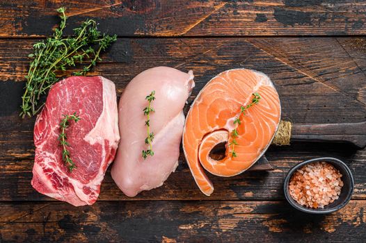 Different types of raw meat steaks Beef striploin, salmon and chicken breast. Dark wooden background. Top view.