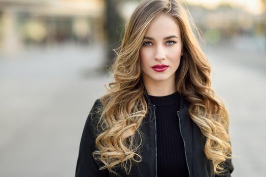 Close-up portrait of young blonde girl with beautiful blue eyes wearing black jacket outdoors. Pretty russian female with long wavy hair hairstyle. Woman in urban background.