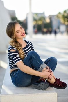 Dreamer blonde woman smiling in urban background. Beautiful young girl wearing striped t-shirt and blue jeans sitting on a bench in the street. Pretty russian female with pigtail and eyes closed.