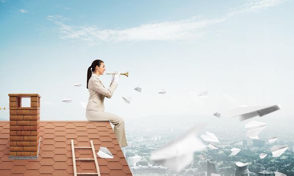 Beautiful young woman playing trumpet on roof. Brave girl in white business suit with music brass instrument sitting on edge of roof. Mixed media business concept. Flying paper planes in blue sky