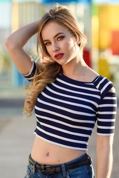 Blonde woman, model of fashion, standing in urban background. Beautiful young girl wearing striped t-shirt and blue jeans in the street. Pretty russian female with pigtail.