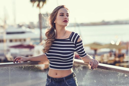 Blonde woman, model of fashion, standing in a harbor with boats and sea in the background. Beautiful young girl wearing striped t-shirt and blue jeans. Pretty russian female with pigtail.