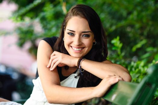 Beautiful smiling young woman with blue eyes looking at camera sitting on urban bench. Happy girl in casual clothes. Lifestyle concept.