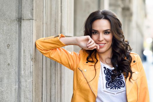 Young brunette woman, model of fashion, wearing orange modern jacket. Pretty caucasian girl with long wavy hairstyle. Thoughtful female in urban background.