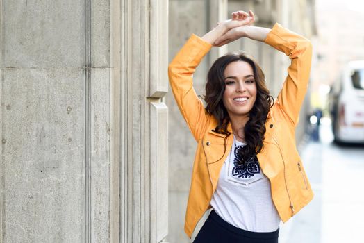Young brunette woman, model of fashion, wearing orange modern jacket and blue skirt. Pretty caucasian girl with long wavy hairstyle smiling. Female raising her arms in urban background.