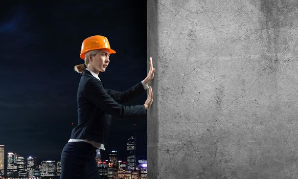 Beautiful woman architect pushing concrete wall. Young specialist in safety helmet on night city background. Overcoming obstacles and solution of problems. Grey concrete wall as banner with copy space