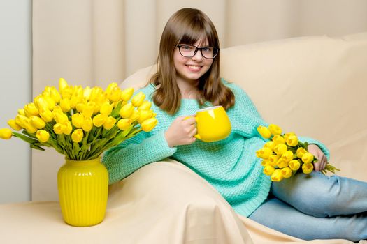 A charming school girl is sitting on the couch with a mug in her hands. Concept of happy people, healthy eating.