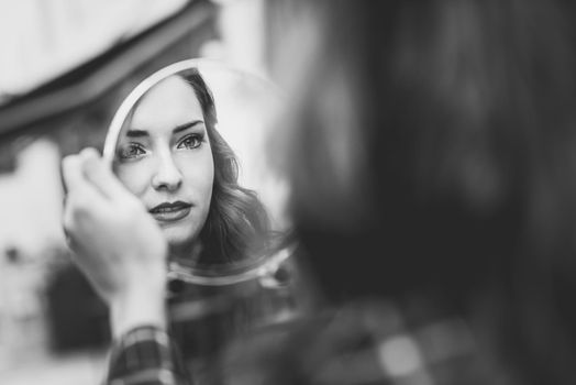 Portrait of young woman looking at herself in a little mirror in urban background. Black and white photograph.