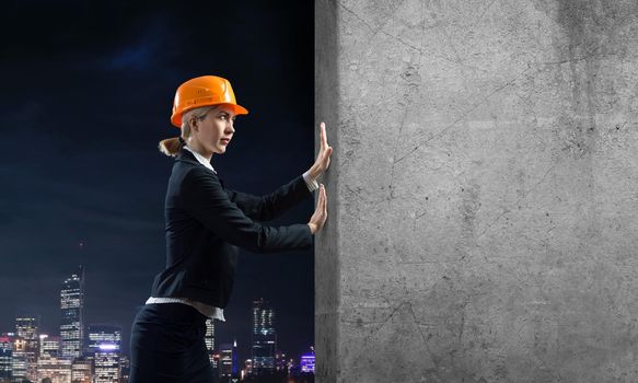 Beautiful woman architect pushing concrete wall. Young specialist in safety helmet on night city background. Overcoming obstacles and solution of problems. Grey concrete wall as banner with copy space