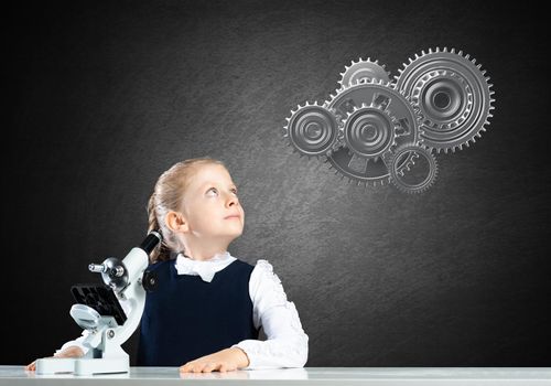 Little girl scientist with microscope on chalkboard background with gears mechanism. Research and discovery concept. Elementary science class in modern school. Schoolgirl in schoolwear sitting at desk