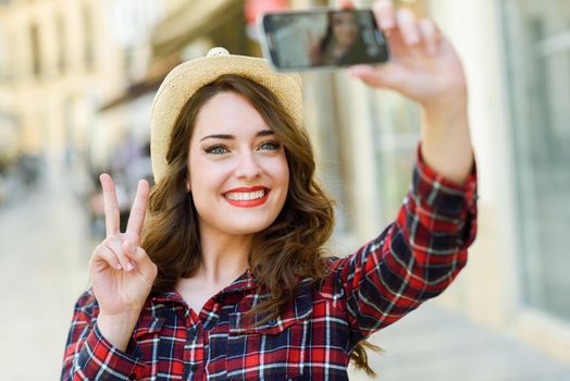 Portrait of a beautiful young woman selfie in the street with a smartphone. Young woman wearing plaid shirt and sun hat with red lips in urban background.