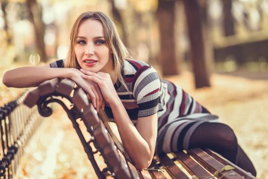 Pensive young blonde woman sitting on a bench in the street of a park with autumn colors. Beautiful girl in urban background wearing striped dress and black tights. Female with straight hair and blue eyes.