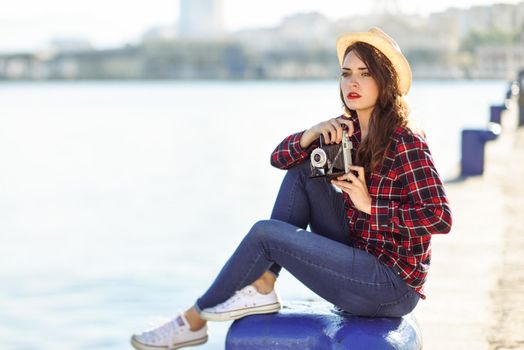 Young woman taking photographs with a vintage camera sitting in a harbour. Girl wearing plaid shirt, blue jeans and sun hat.