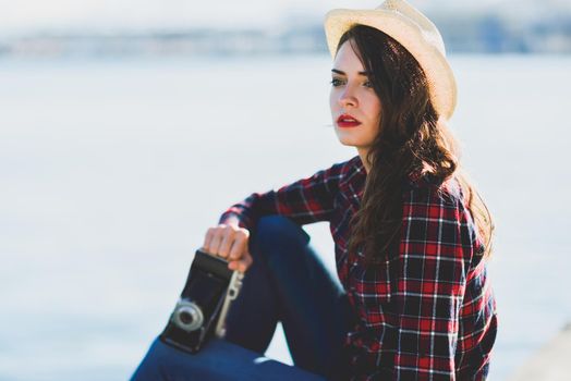 Young woman taking photographs with a vintage camera sitting in a harbour. Girl wearing plaid shirt, blue jeans and sun hat.