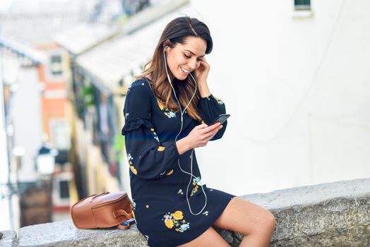 Woman listening to the music with earphones and smart phone in urban background. Female in casual clothes with care hairstyle in Granada, Andalusia, Spain.