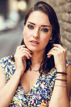 Beautiful young woman with blue eyes standing next to brick wall outdoors. Girl wearing flower dress in urban background. Beauty and fashion concept.