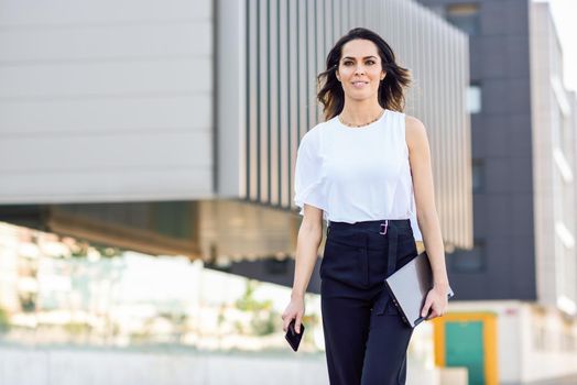 Young businesswoman working with her smart phone and laptop outdoors. Middle-aged woman walking near business building with very careful hair.