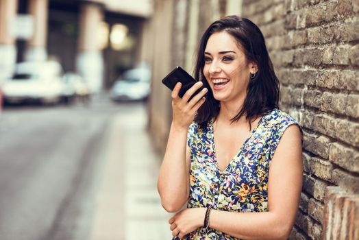 Smiling young woman recording voice note in her smart phone outdoors. Girl wearing flower dress in urban background. Technology concept.