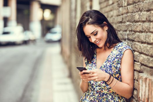 Smiling young woman using her smart phone outdoors. Girl wearing flower dress in urban background. Technology concept.