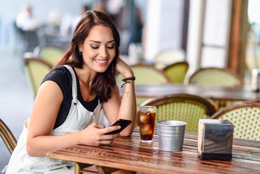 Smiling young woman with blue eyes sitting on urban cafe using smart phone. Girl with brown wavy hairstyle wearing white denim dress. Lifestyle concept.