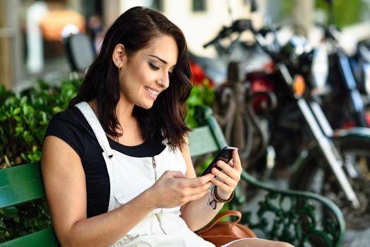 Smiling young woman looking at smart phone outdoors. Girl wearing white denim dress sitting in urban bench. Technology concept.