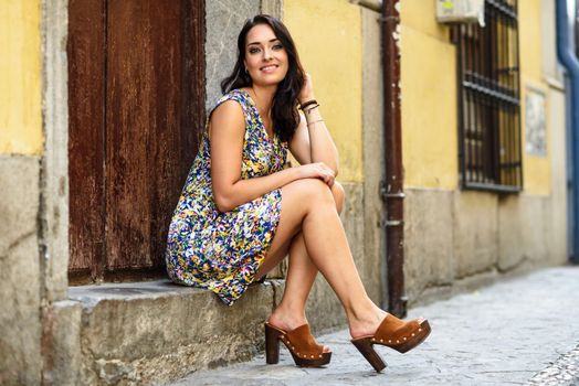 Happy young woman with blue eyes smiling sitting on urban step. Girl wearing flower dress in urban background. Beauty and fashion concept.
