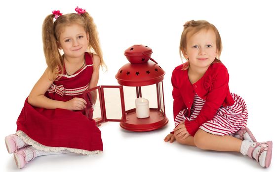 Two beautiful little girls sisters dressed in red dresses . girl sitting on the floor near the lantern in which the candle burns - Isolated on white background