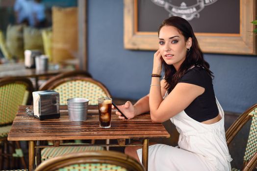 Beautiful girl with blue eyes sitting on urban cafe using smart phone. Young woman with brown wavy hairstyle wearing white denim dress. Lifestyle concept.