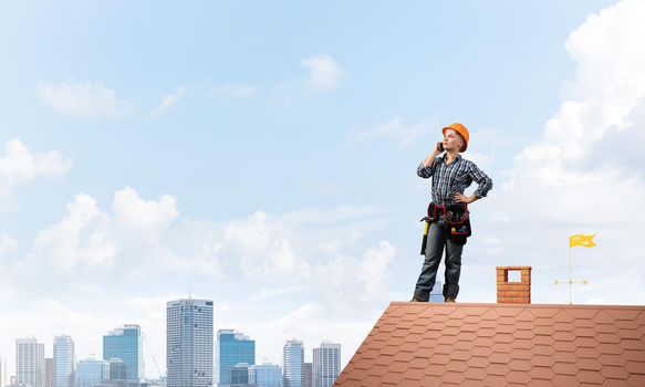Attractive blonde woman in workwear and hardhat talking on smartphone. Young worker in checkered blue shirt standing on brick roof of building. Professional renovation and roofing construction.