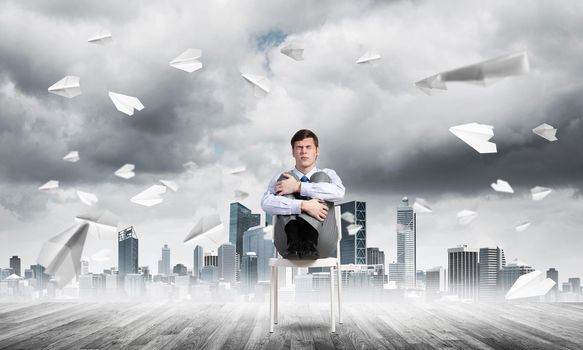 scared businessman sitting on a chair with his hands clasped his knees, paper plane fly around
