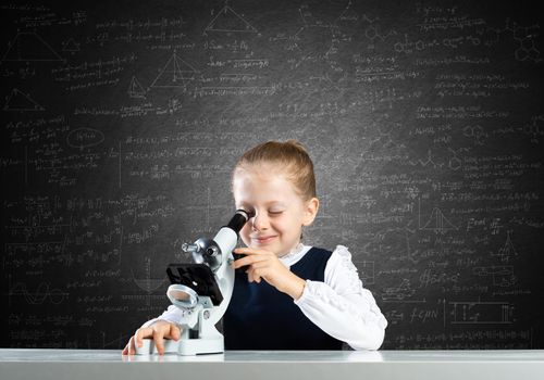 Little girl scientist looking through microscope in classroom at lesson. Research and discovery. Formal education in modern school. Schoolgirl in schoolwear making experiment in class with chalkboard.
