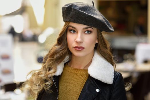 Blonde russian woman in urban background. Beautiful young girl wearing beret, black leather jacket and mini skirt standing in the street. Pretty female with long wavy hair hairstyle and blue eyes.