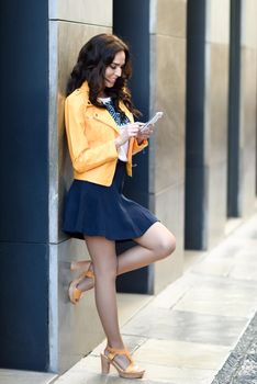 Young brunette woman, model of fashion, wearing orange modern jacket and blue skirt, texting with her smart phone in urban background.