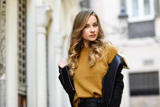 Blonde woman in urban background. Beautiful young girl wearing black leather jacket and mini skirt standing in the street. Pretty russian female with long wavy hair hairstyle and blue eyes.