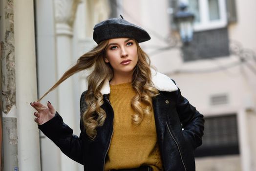 Blonde russian woman in urban background. Beautiful young girl wearing beret, black leather jacket and mini skirt standing in the street. Pretty female with long wavy hair hairstyle and blue eyes.