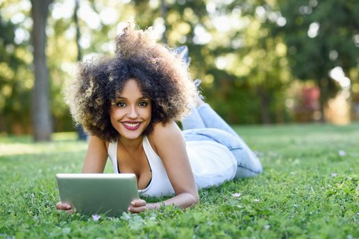 Young black woman with afro hairstyle laying on grass in urban park looking at camera and playing with her tablet computer. Mixed girl wearing casual clothes.