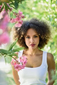 Young black woman with afro hairstyle in urban park. Mixed girl wearing casual clothes between pink flowers.