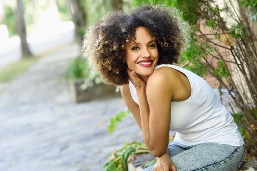 Young black woman with afro hairstyle sitting and smiling in urban park. Mixed girl wearing white t-shirt and blue jeans.