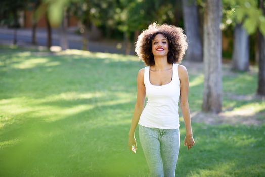 Young black woman with afro hairstyle smiling in urban park. Mixed girl wearing white t-shirt and blue jeans walking on the grass.