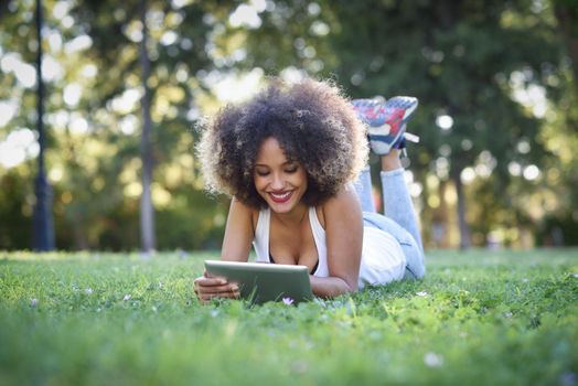 Young black woman with afro hairstyle laying on grass in urban park looking at her tablet computer. Mixed girl wearing casual clothes.
