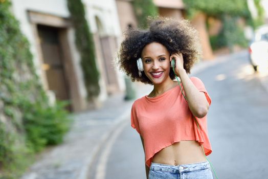 Portrait of young attractive black girl in urban background listening to the music with headphones. Woman wearing leather jacket and blue jeans with afro hairstyle