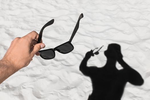 The young man's hand holds sunglasses against the background of his black silhouette shadow on the white beach sand.